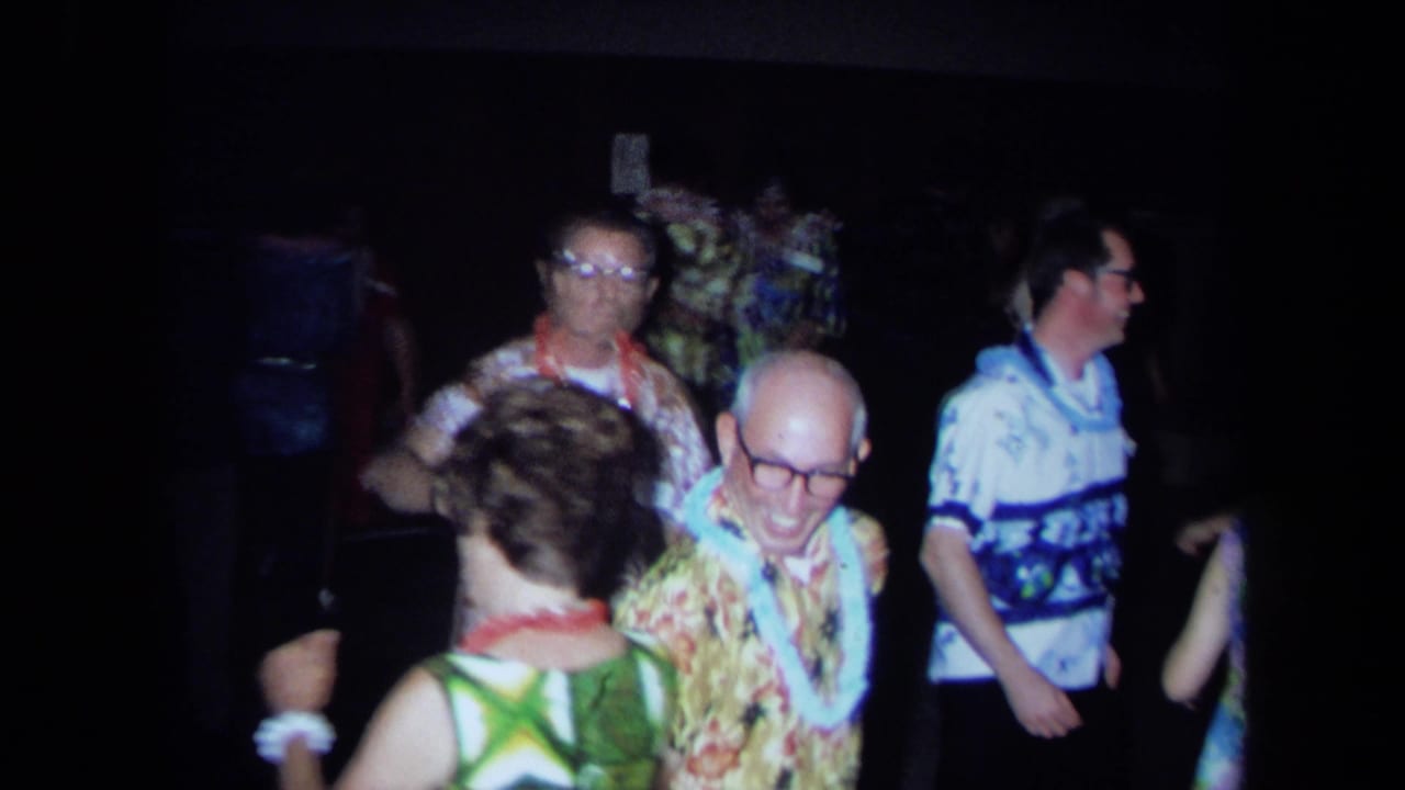 Adults in leis and Hawaiian-style shirts dance and smile in a dark hall.