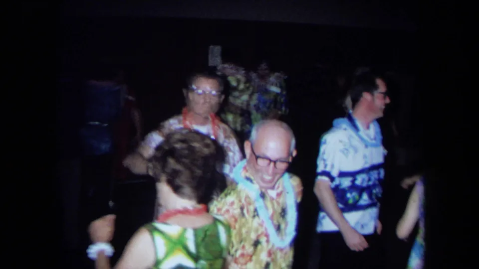 1971 California party scene with adults wearing leis, floral clothing, and patterned shirts on a dim dance floor.