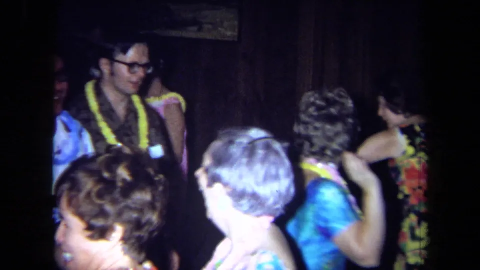 1971 California home-movie frame of adults in leis and colorful party clothes dancing in a dark indoor space.