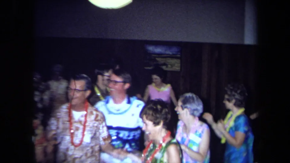The frame shows a small group at the 1971 California Hawaiian-themed party, wearing leis and aloha-style clothing under dim lighting.