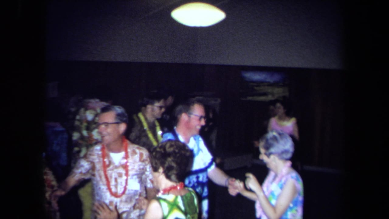 Adults at a 1971 California Hawaiian-theme party wear leis and patterned clothing while dancing in a dim indoor room.