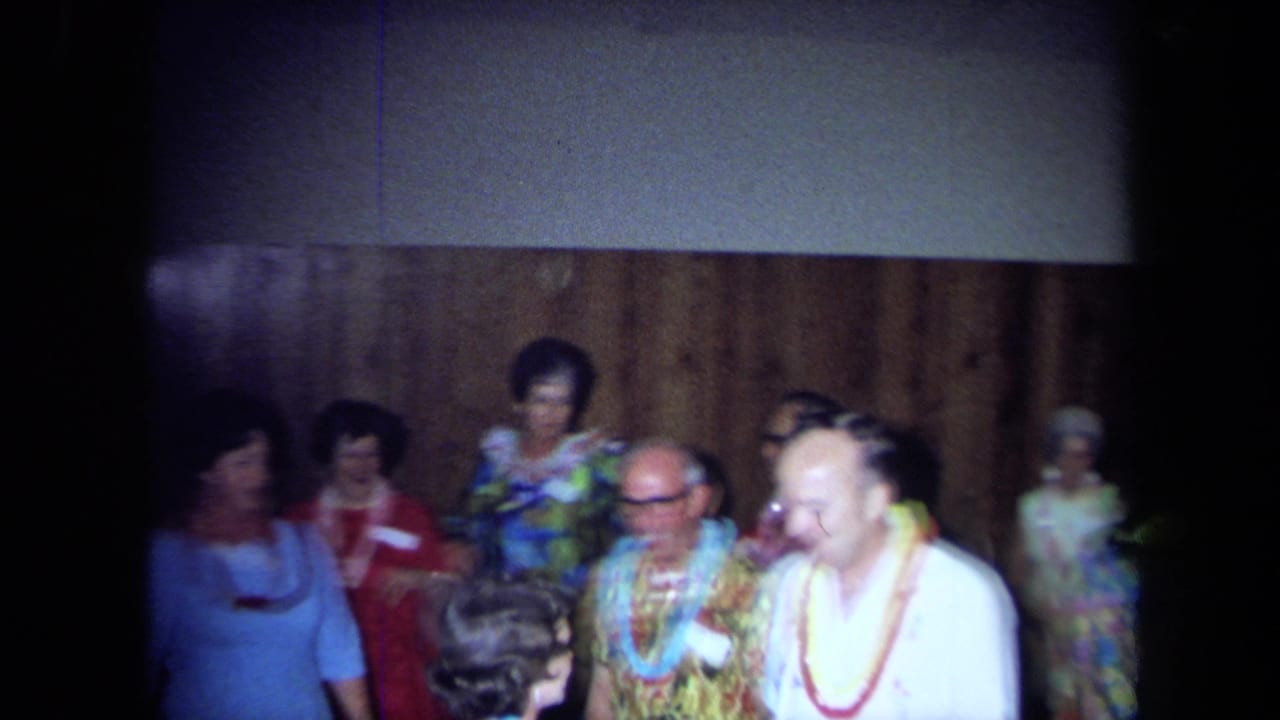 Party guests in leis stand and talk near a wood-paneled wall.