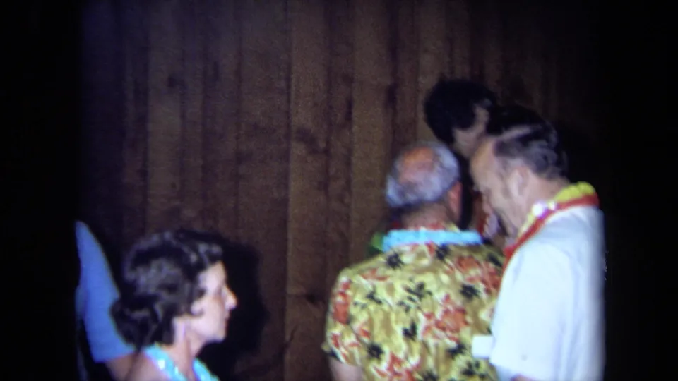 Adults wearing leis converse in a dim wood-paneled interior at a California home-movie party from 1971.
