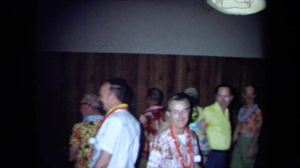 Adults wearing leis and colorful shirts mingle in a 1971 California Hawaiian-theme party scene filmed on 8mm.