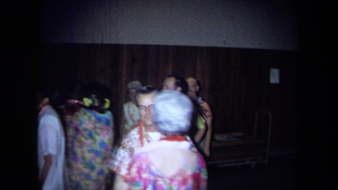 1971 California home-movie frame showing adults in leis, aloha-style clothing, and floral dresses mingling in a dark interior.
