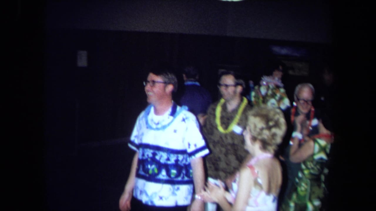 Smiling adults wearing leis stand and dance in a dark party room.
