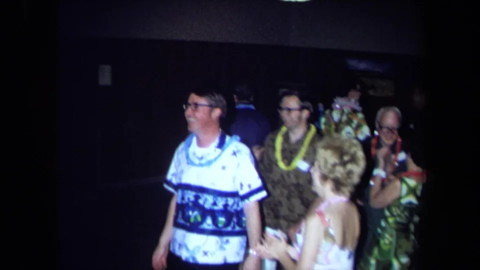 Adults at a 1971 California party wear leis and patterned shirts while standing and dancing in low indoor light.