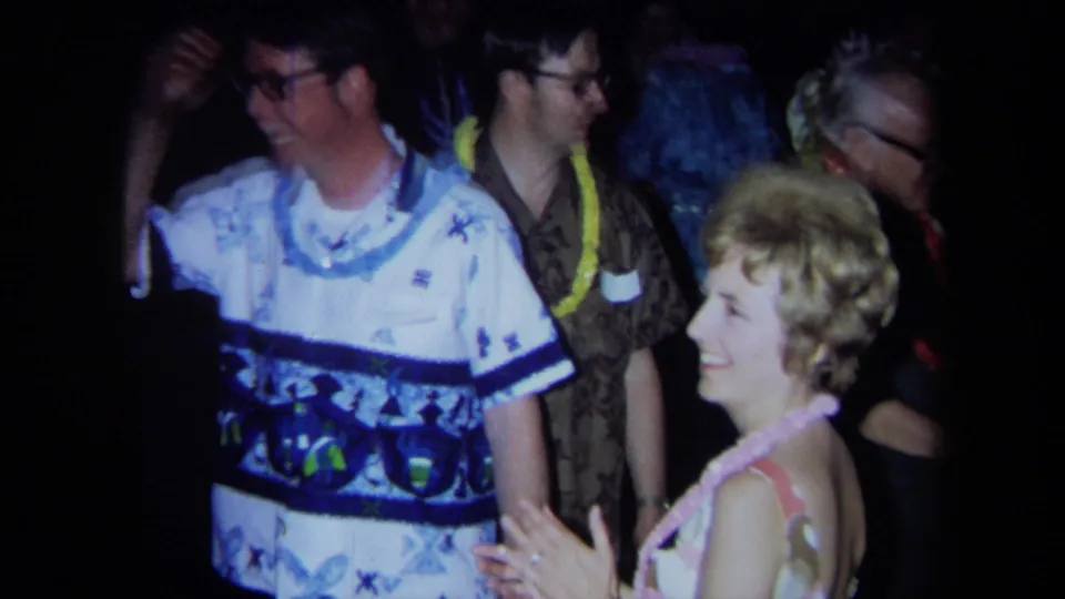 Close home-movie view of adults in leis and patterned clothing laughing and dancing at a 1971 California indoor party.