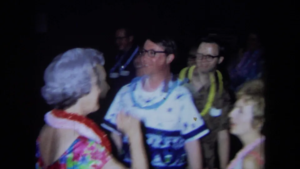 1971 California home-movie frame of adults in leis and aloha-style clothing dancing and smiling in a dark indoor party space.