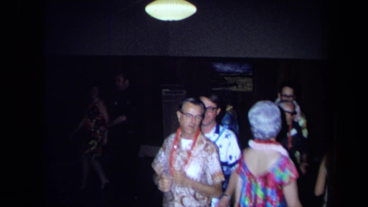 Adults in aloha-style clothing and leis dance indoors at a California home-movie luau scene from the supplied 1971 metadata.