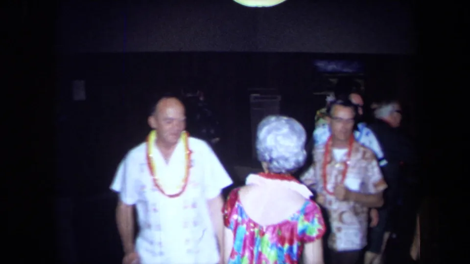At the 1971 California luau party, a man in a white shirt and lei stands near a woman in a bright dress as guests dance in the background.