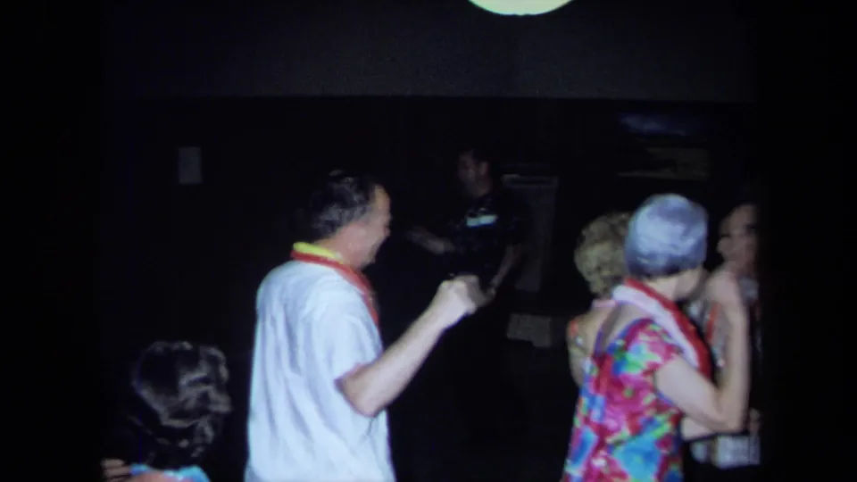 Adults in leis and party clothing dance and mingle in a dim California interior, consistent with the supplied 1971 luau home-movie metadata.
