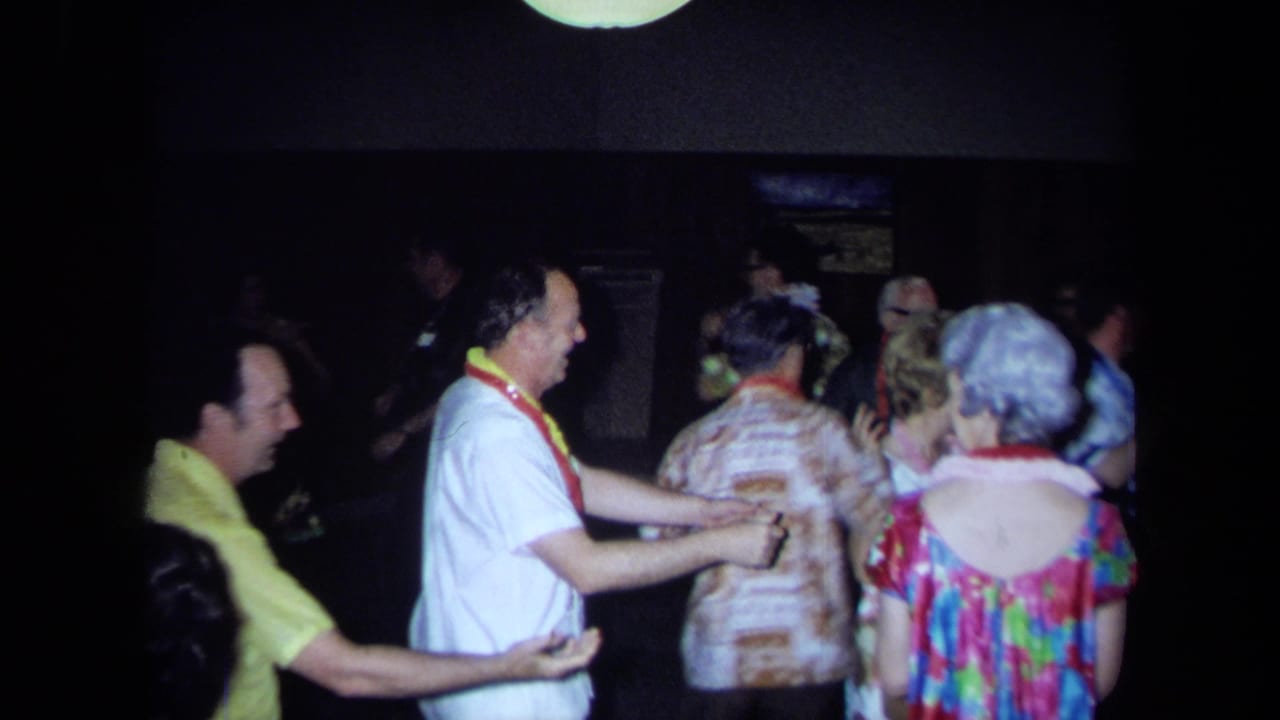 California 1971 home-movie frame of adult partygoers in Hawaiian print clothing and leis dancing in a dark indoor space.