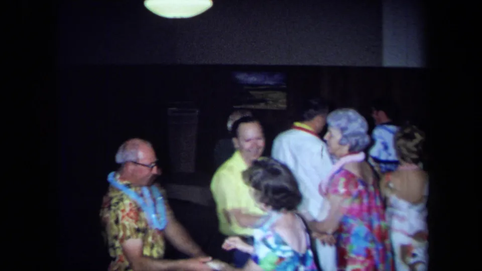 1971 California home-movie frame showing adults in leis and patterned shirts dancing or mingling in a dim private interior.
