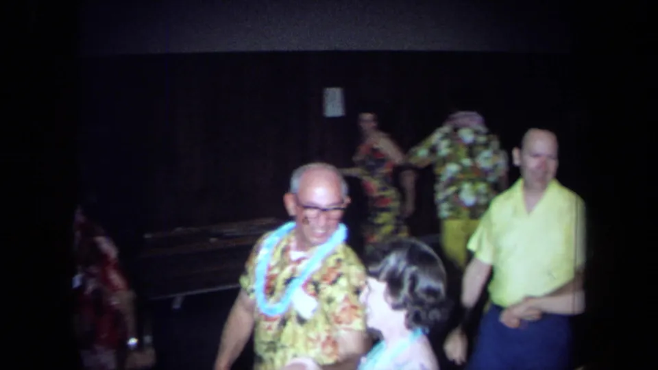 Adult man in a Hawaiian print shirt and lei dances near other partygoers in a dim California indoor setting from 1971 metadata.