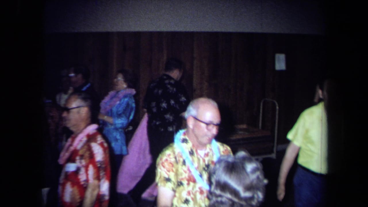 1971 California home-movie frame showing adults at a Hawaiian-themed indoor party with leis, aloha shirts, and wood-paneled walls.