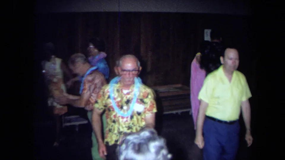 1971 California party frame with a man in a yellow floral shirt and blue lei among adults in a dim private interior.