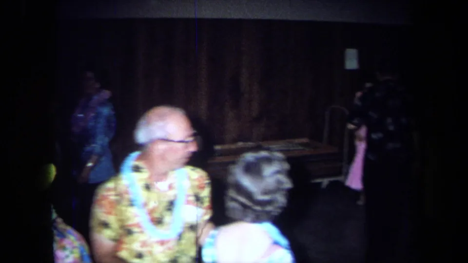 Handheld 1971 California home-movie frame showing adults moving and mingling at a Hawaiian-themed indoor party.