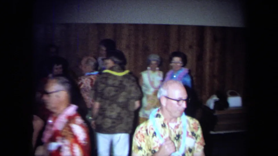 1971 California home-movie frame showing adults in leis and patterned clothing gathered in a dim party room.