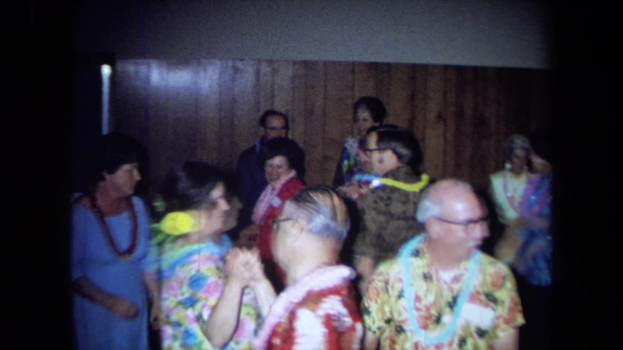 Older guests in leis and floral clothing gather and dance in a wood-paneled room.