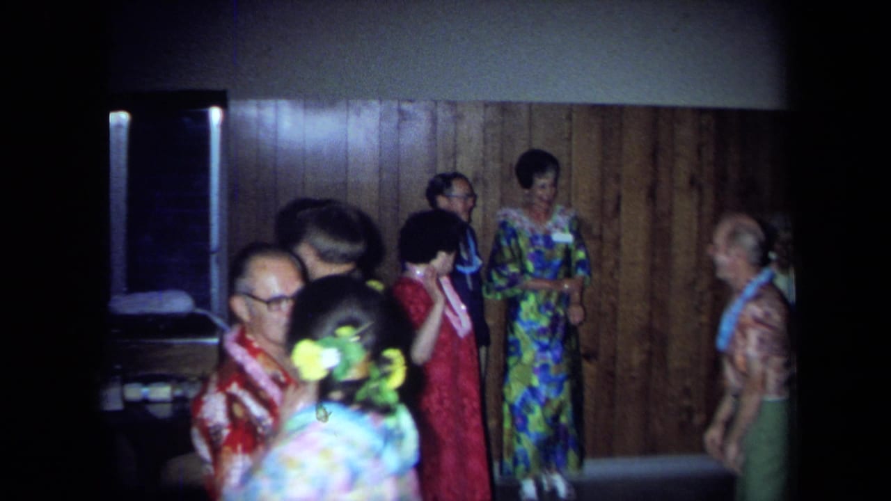 Party guests in colorful clothing stand near a wood-paneled wall and dark opening.