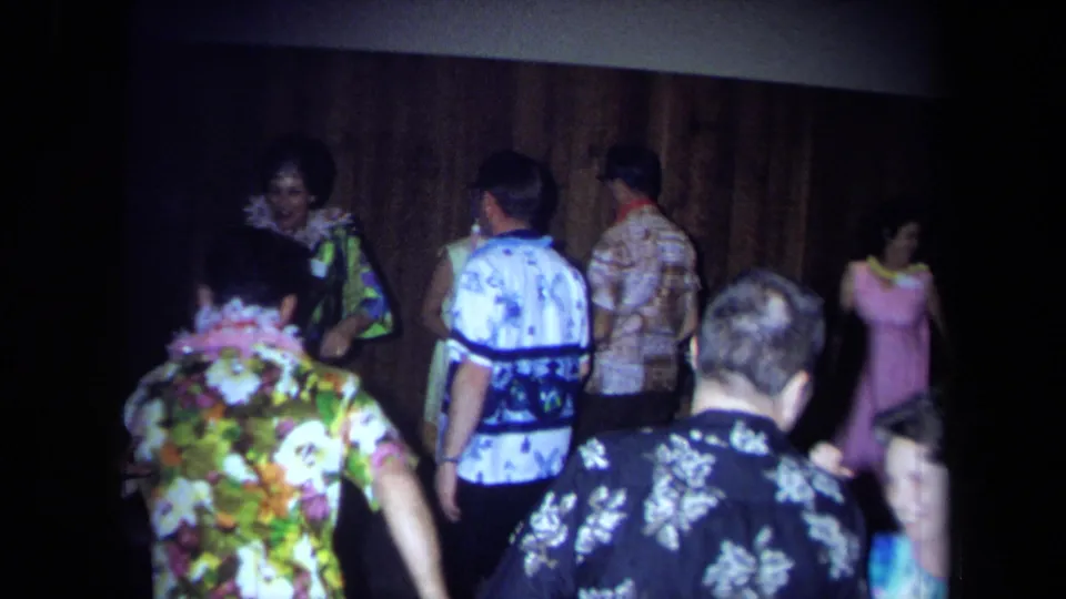 Several older guests in floral shirts and leis move through the wood-paneled party room during the 1971 California home movie.