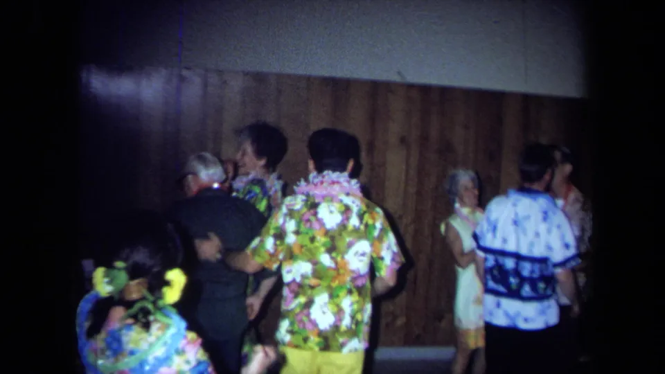 Older guests in bright floral clothing and leis dance and mingle inside a private California party room in 1971.
