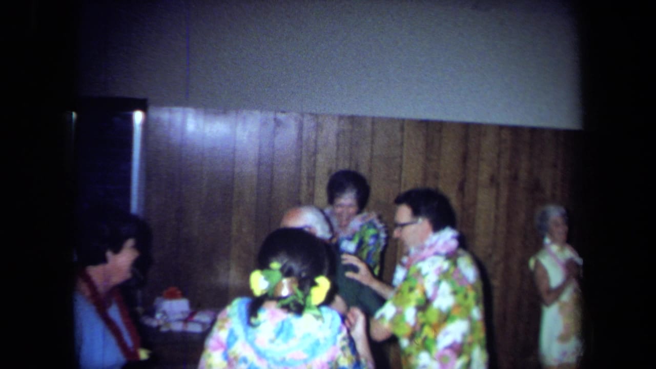 1971 California home-movie frame showing adults in leis, aloha-style clothing and floral dress near a wood-paneled wall.