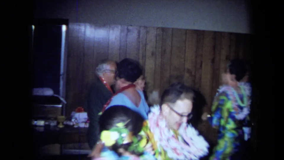 Close 8mm frame of adults in leis and patterned party clothes moving through a private 1971 California interior.