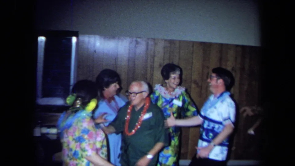 Group of adult party guests in leis, floral clothing and aloha-style shirts inside a 1971 California home-movie scene.