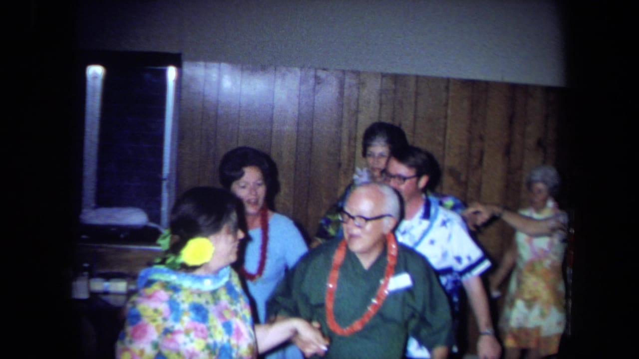 Adults at a 1971 California indoor party wear leis and patterned clothing while dancing in front of wood-paneled walls.