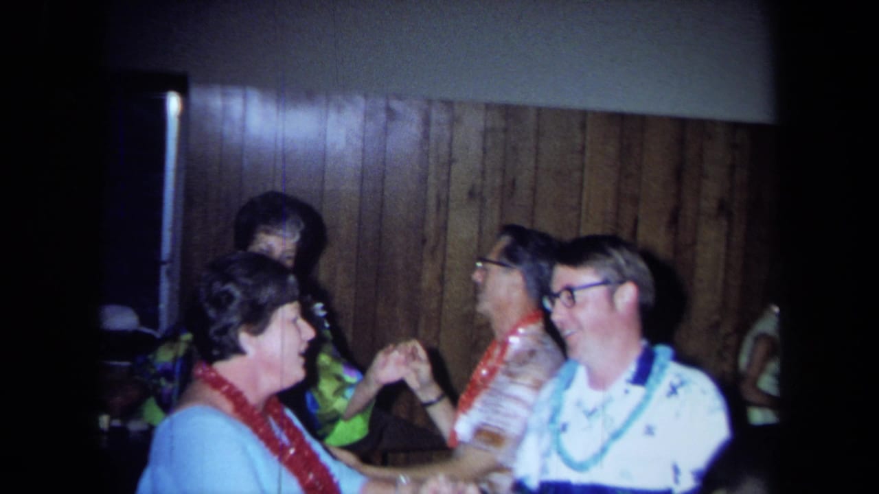 Adults in leis dance and hold hands beside a wood-paneled wall.