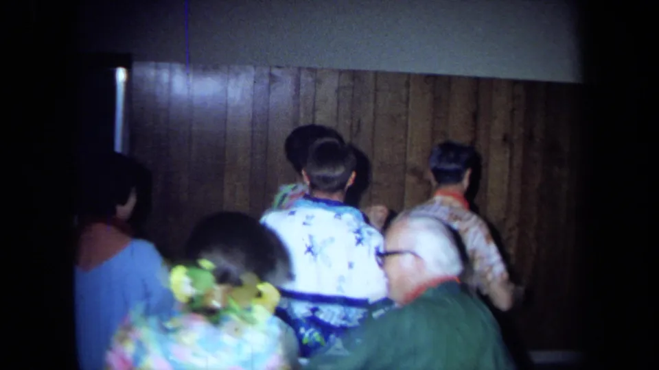 A 1971 California home-movie frame shows adults from behind as they dance in leis and patterned shirts inside a private room.