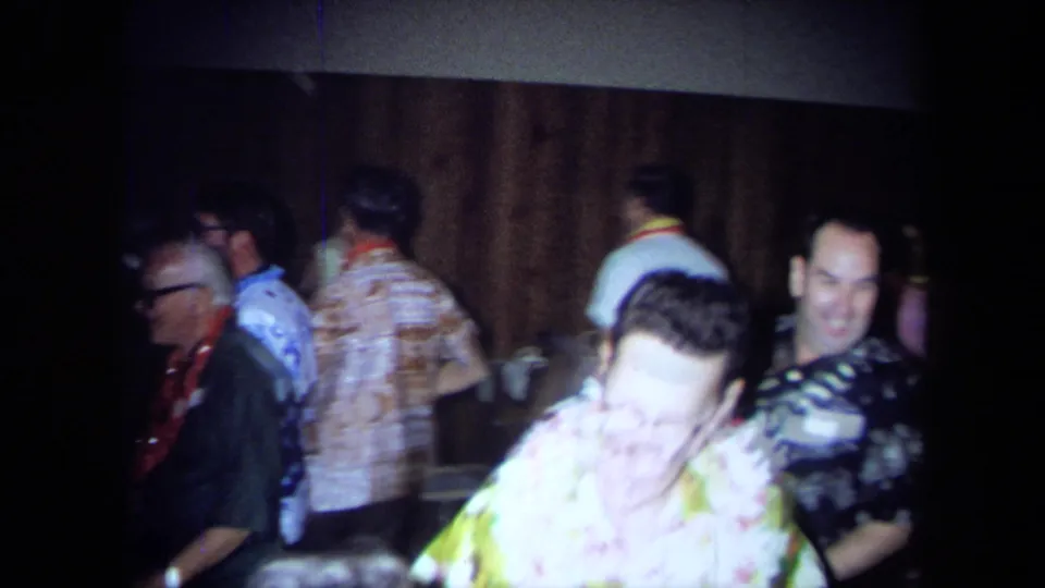 Adults at a 1971 California indoor party move past the camera in leis and bold patterned shirts, with soft focus and warm room light.