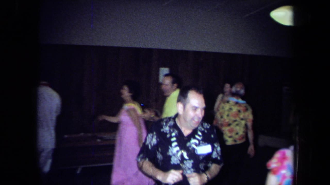 1971 California home-movie frame of adults dancing indoors, with leis, patterned shirts, wood paneling, and a handheld 8mm look.