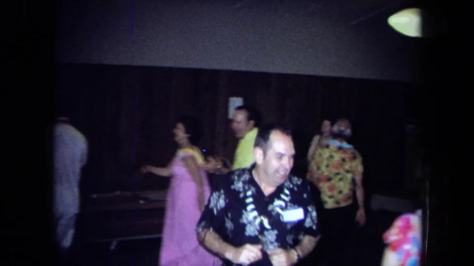 1971 California home-movie frame of adults dancing indoors, with leis, patterned shirts, wood paneling, and a handheld 8mm look.