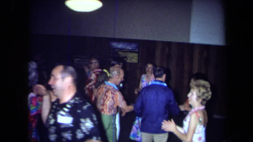 Adults in leis and colorful clothing gather and dance inside a dim California room in this 1971 8mm home-movie frame.