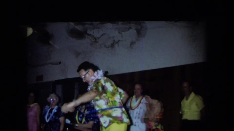 A man in glasses and a bright floral shirt hula-hoops in a dim indoor California party scene from 1971, with other adults watching.