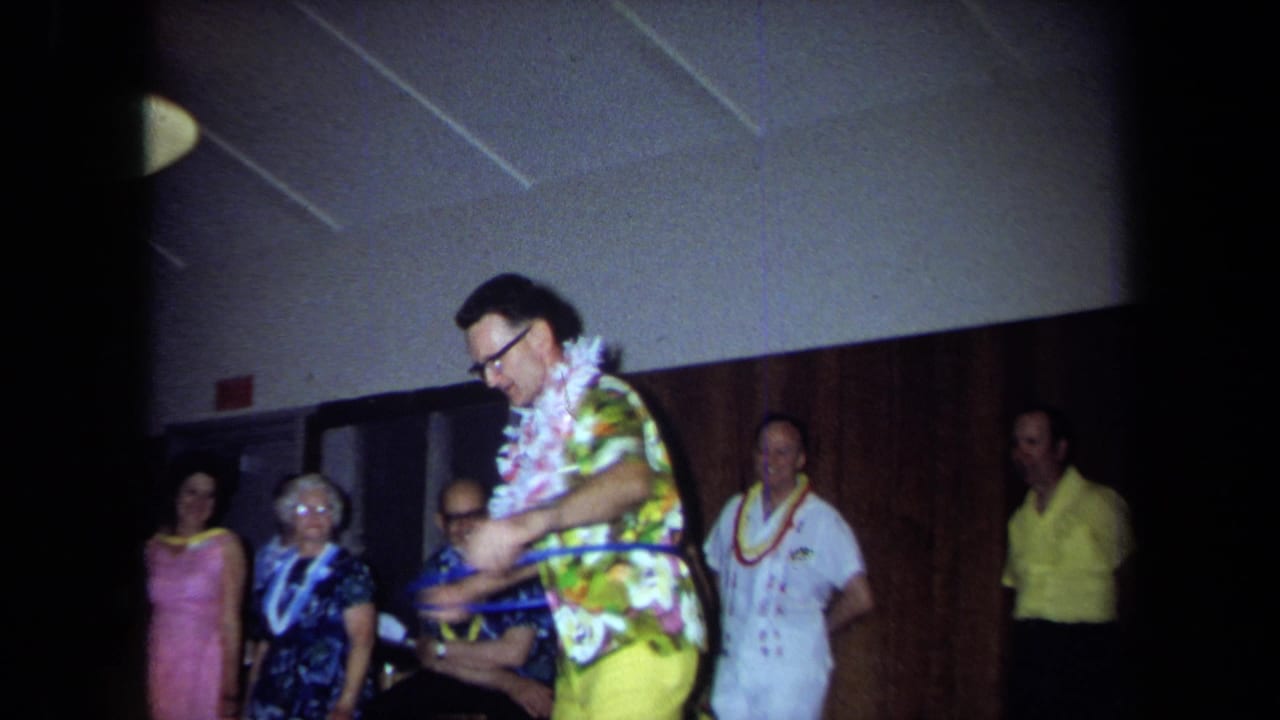 In 1971 California home-movie footage, a man wearing a lei, glasses, floral shirt, and yellow pants hula-hoops before party guests.