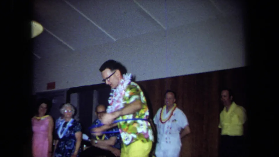 In 1971 California home-movie footage, a man wearing a lei, glasses, floral shirt, and yellow pants hula-hoops before party guests.