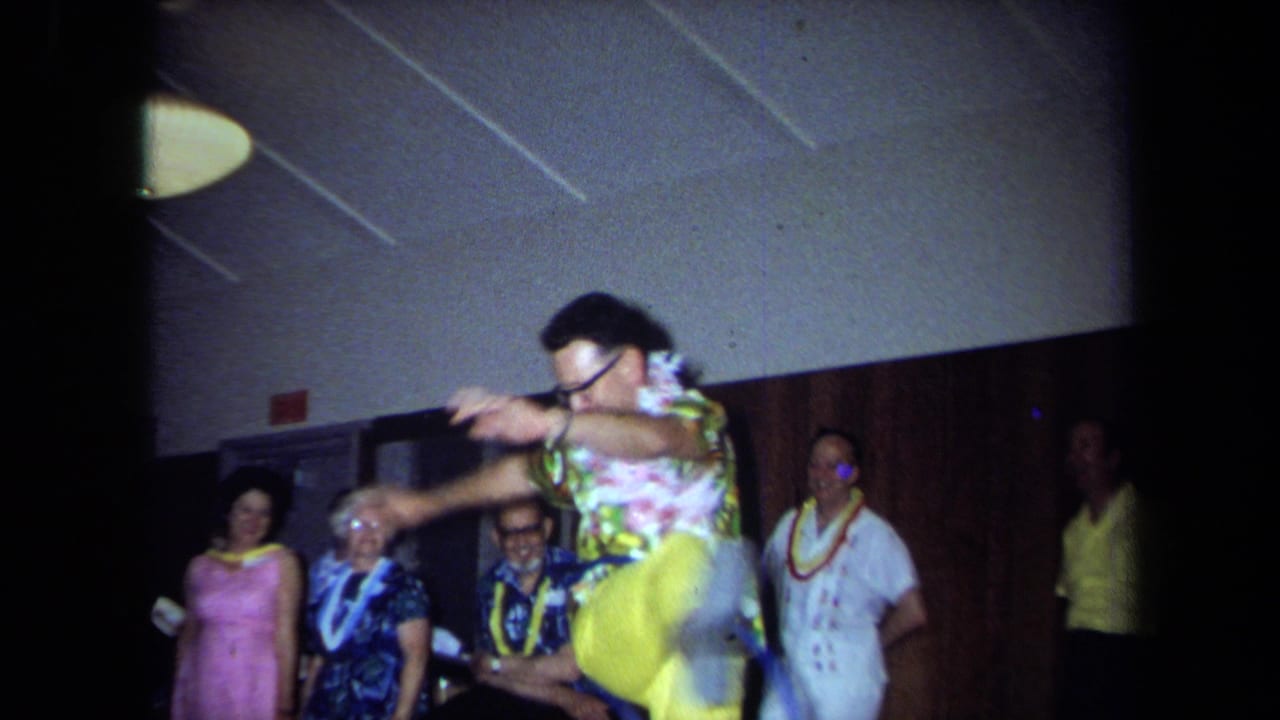 Man raises a knee while hula-hooping in front of guests wearing leis and bright clothing.