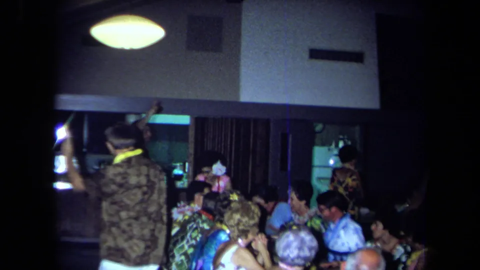 A crowded 1971 California indoor gathering shows seated adults at tables while a standing guest raises an arm in the party room.