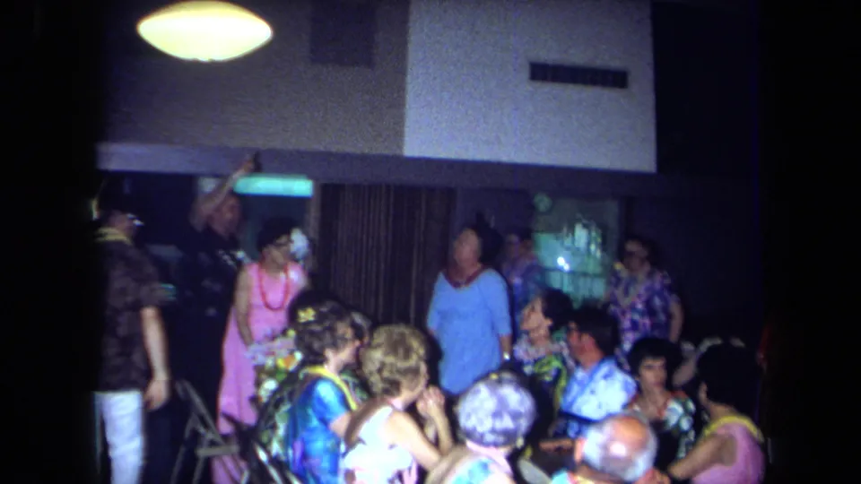 The 1971 California home movie shows adults standing and sitting in a lively private interior with leis, bright clothing, and party tables.