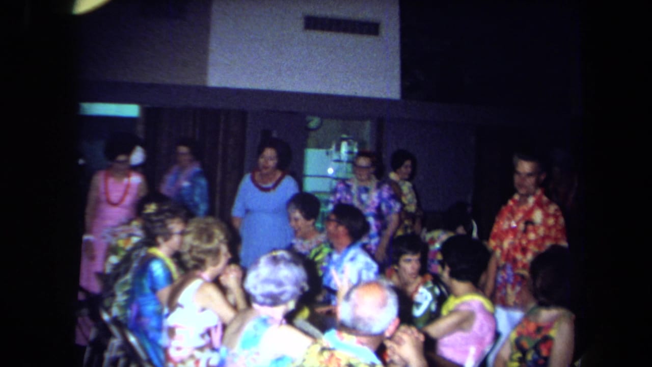 1971 California home-movie frame showing adults in bright shirts, dresses, and leis gathered in a dim private party room.