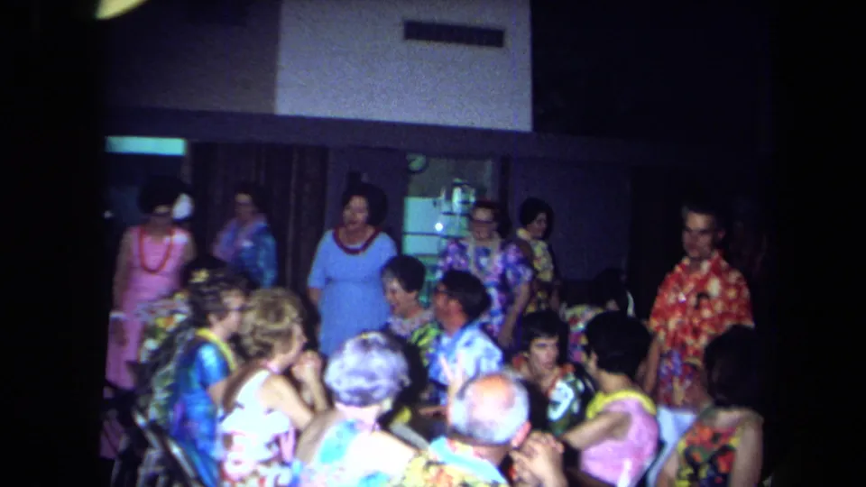1971 California home-movie frame showing adults in bright shirts, dresses, and leis gathered in a dim private party room.