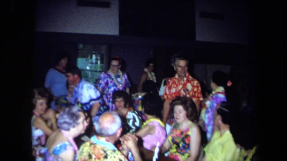 1971 California party scene with adults in bright patterned clothing and leis seated and standing in a crowded interior.