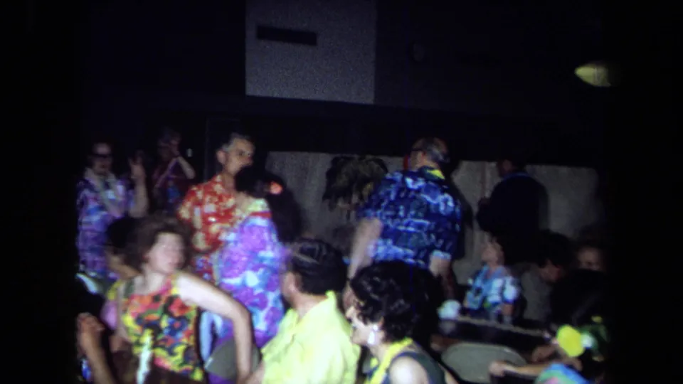 Handheld 8mm frame from a 1971 California indoor party, with adults in leis and patterned shirts moving through the room.