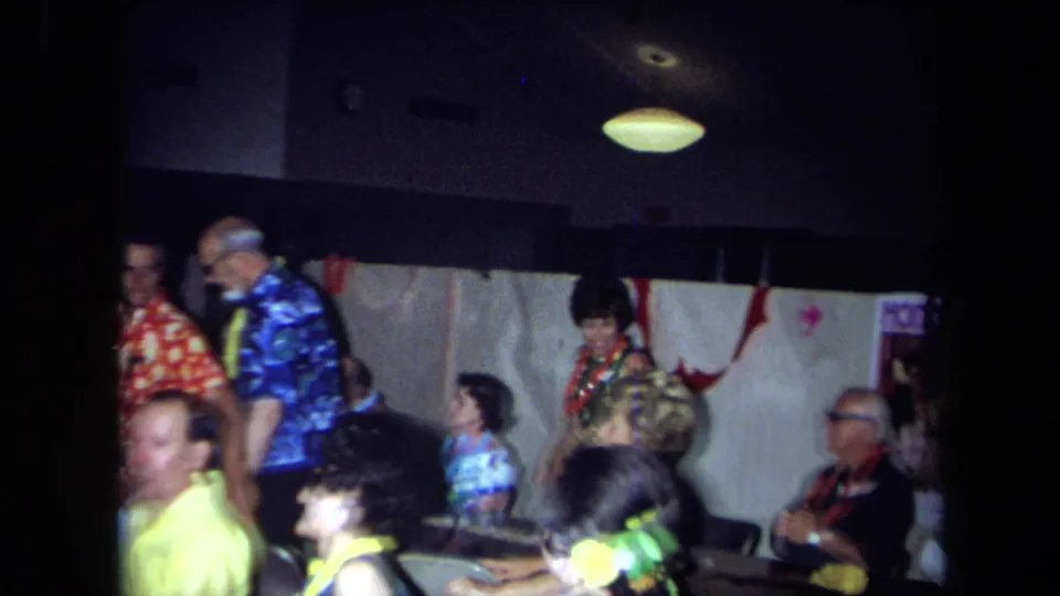 1971 California home-movie party frame with adults in Hawaiian-style shirts and leis near wall decorations and tables.