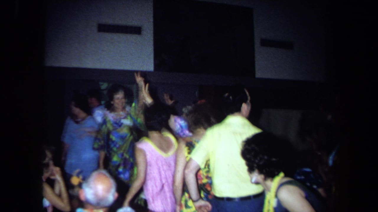 Adults in leis and tropical clothing dance in a dim indoor hall.