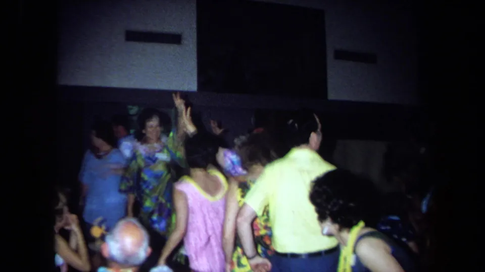 1971 California home-movie frame showing adults at a Hawaiian-theme reunion dancing in a dim hall with leis and bright clothing.
